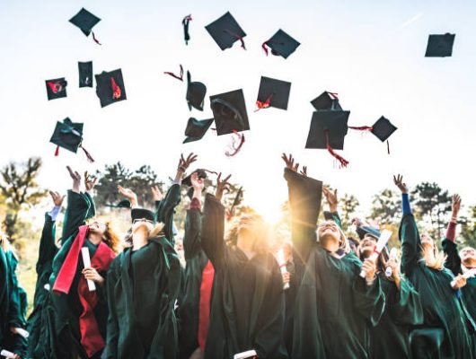 Large group of happy college students celebrating their graduation day outdoors while throwing their caps up in the air.